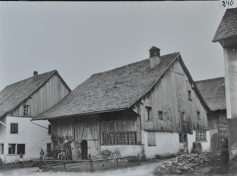 Mehrstöckiges Wohnhaus mit gemauerten Fundament, oberer Teil aus Holz, Satteldach mit Kamin, Scheunentor offen, daneben liegt ein Holzkarren, mehrere Personen posieren, ein Weg führt vor und neben das Gebäude vorbei
