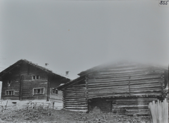 Dreistöckiges Bauernhaus mit Fundament aus Stein, obere Etagen aus Holz, Satteldach mit Kaminen, Garten mit Gartenzaun vor dem Haus, rechts vom Wohnhaus befindet sich Scheune aus Holz, davor gestapelte Holzbalken
