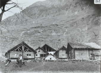 Ansicht eines Dorfteils mit mehrereen Gebäuden aus Holz und Fundament aus Stein, Satteldächer, vor den Häusern verläuft eine Steinmauer, davor Kreuz im Boden, im Vordergrund posiern mehrere Männer, teils sitzend