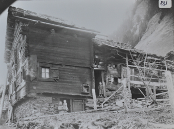 Mehrstöckiges Bauernhaus mit Fundament aus Stein, oberr teils aus Holz, Satteldach, weiterer Anbaus aus Holz und Stein, vor der Türe posiert Frau mit Kind im Arm, wetere Bube posiert auf Steintreppe, Berg im Hintergrund