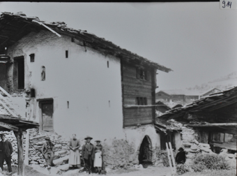 Zweistöckiges Wohnhaus mit gemauerten Fundament und Wànden, vorderer Teil der Wand aus Holzbau, Satteldach, auf der Steintreppe posieren mehrere Personen, Heiligenfigur in die Fassade oberhalb des Eingangs eingebaut, im Hintergrund weitere Gebäude
