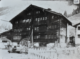 Mehrstöckiges Mehrfach-Wohnhaus aus Holz mit Fundament aus Stein, Satteldach mit Kamin, Brennholz vor dem Haus, auf dem Vorplatz Brunnen, wo eine Frau und zwei Kinder posieren, Gartenzaun im Vordergrund, weitere Gebäude in Umgebung