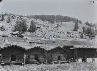 Ansicht von drei Holzhütten des Dorfes, Hütten stehen auf Pfeilern, Satteldächer, rechts neben den Hütten steht ein Wohnhaus aus Holz mit gemauertem Fundament, Strasse vor den Hütten, Hügel im Hintergrund
