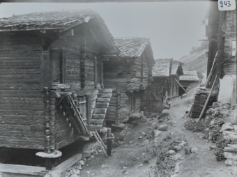Ansicht eines Dorfteils mit mehreren Häusern, Häuser sind aus Holz und mit Satteldach, die Häuser stehen auf Sockeln, vor dem ersten Haus posiert ein Bube, ein Feldweg führt durch das Dorf