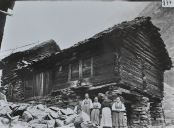 Wohnhaus mit Wänden aus Holz, Fundament aus Stein, Haus steht am vorderen Ende auf Pfeilern aus Stein, Satteldach, fünf Personen posieren vor dem Haus, am hinteren Ende des Hauses ist ein weiterer Anbaus aus Holz und Stein