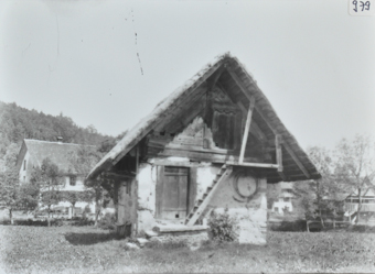 Zweistöckiges Wohnhaus, unterer Teil gemauert, oberer Teil aus Holz, Schleppdach, Holztreppe fürht zum oberen Geschoss, Haus steht auf Wiese, im Hintergrund Bäume sowie weitere Häuser
