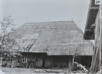 Mehrstöckiges Bauernhaus aus Holz, grosses Walmdach bzw. Reetdach, Brennholz sowie weitere Gegenstände liegen vor dem Haus, zwei Bäume stehen vor dem Haus