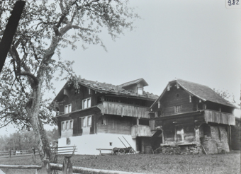 Ansicht von zwei Bauernhäuser aus Holz und Fundament aus Stein, Satteldach resp. Satteldach mit  Gaube, Holzkarren steht vor dem Haus, vor den Häusern ist eine Wiesen, im Vordergrund steht ein Baum