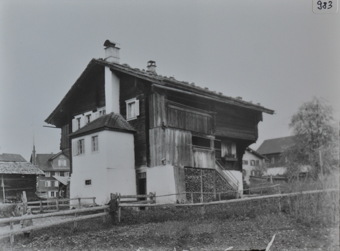 Mehrstöckiges Bauernhaus, gemauertes Fundament, Wände aus Holz, Satteldach mit Kaminen, Treppe führt zum Hauseingang auf der oberen Etage, unter der Treppe liegt gestapeltes Brennholz, vor dem Haus ist gemauerter Anbau, vor dem Haus durchläuft Strasse, vor der Strasse posieren zwei Kinder an einem Holzzaun