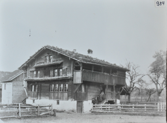 Dreistöckiges Bauernhaus mit Wänden aus Holz, gemauertes Fundament, Satteldach und Balkon, auf der Treppe posieren mehrere Personen, Strasse vor dem Haus, hinter dem Haus Wiese mit Bäumen