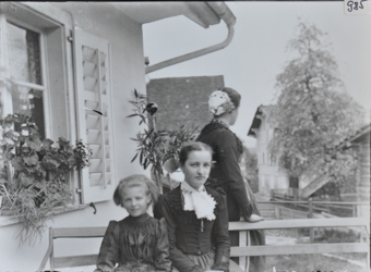 Drei Frauen posieren vor einem Haus, davon zwei sitzend auf einer Bank, Blumen auf dem Fenstersims