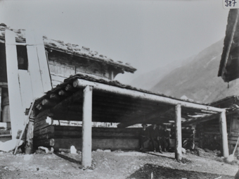 Vier Männder posieren sitzend unter einem Unterschlupf aus Holz, das Dach ist mit Holzpfeilern gestützt, im Hintergrund weiteres Gebäude