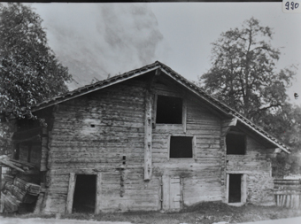 Mehrstöckige Scheune aus Holz, Schleppdach, Öffnungen in den Wänden ohne Fenster, links an der Wand steht gestapeltes Holz, belaubter Baum links am Haus