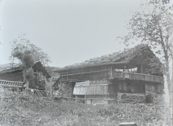 Zweistöckiges Bauernhaus aus Holz, Satteldach, Wäsche hängt auf einer Leine auf dem Balkon, mehrere Stapel Brennholz vor und neben dem Haus, lange Holztreppe ist quer and die Wand befestigt, auf der Strasse vor dem Haus posieren zwei Kinder, davor Brunnen