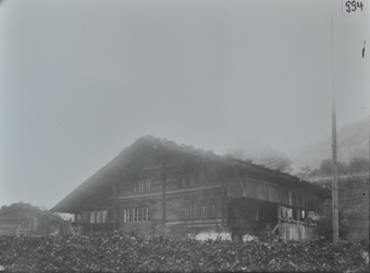 Zweistöckiges Bauernhaus mit Wänden aus Holz und gemauerten Fundament, Schleppdach, aufgehängte Wäsche unter dem Balkon, rechts steht weiteres Gebäude aus Holz