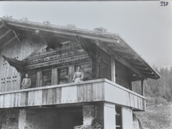 Zwei Frauen posieren auf einem Balkon aus Holz, Gebäude aus Holz mit Satteldach, Haus steht auf Pfeilern aus Stein, Fassade mit Bildern und einer Inschrift