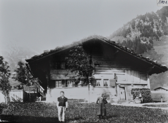 Zweistöckiges Bauernhaus aus Holz, gosses Satteldach, gestapeltes Brennholz rechts an der Hauswand, Baum vor dem Haus, vor dem Haus Wiese mit zwei posierenden Kindern, Hügel im Hintergrund