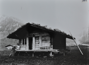Einstöckiges Wohnhaus aus Holz mit Satteldach, Dach mit Steinen verstärkt, Haus steht auf Holzpfeilern, Leiter rechts an die Wand angelehnt, vor dem Haus posieren zwei Frauen, Brennholz unter dem Haus