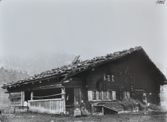 Zweistöckiges Bauernhaus aus Holz, Schleppdach mit Steinen verstärkt, zwei Frauen posieren sitzend vor dem Haus, Brennholz vor dem Haus