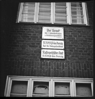 DANZIG: Officially, a "free town" with the Senate an independant government | under Polish protection this picture proves otherwise...the rule of the | German Nazi party. | [Einzug] The first sign is for the Senate's Department for Public Health, the | second and third are for the German National Socialist Party's departments | for public health and racial politics. In fact, the three departments form | a single organization, occupying the same o ffice [sic! office]. | [Einzug] This illustrates the absurd situation of the Danzig government; the | President of the Senate, Greiser, ranging second in the [durchgestrichenes Wort: Nazi] Danzig-Nazi | party. Forster, the leader of of [sic!] the Nazis in Danzig being the real ruler | of the "Free Town." [sic! "free town".] | [maschinengeschriebener Eintrag auf separatem Beiblatt; ohne Titel]The Senat of the Free Town of Danzig is supposed to be the real govern- | ment. But the President of the Senat-is [sic!] only the second man in the | Nazi Party (NSDAP), which is an absurd situation. Jt [sic! It] is illustrated by | one of the pictures, showing the Health Department of the Senat and the | Department for Racial Politic of the Nazi Party beeing [sic! being] the same and | one office. [...] | [maschinengeschriebener Eintrag auf separatem Beiblatt; Titel: "Photos Annemarie Clark-Schwarzenbach / Danzig 1937 - Gdynia"; versehen mit Stempel: Credit: / from BLACKSTAR / GRAYBAR BUILDING NEW YORK)]