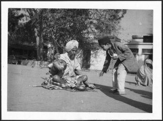 Britisch-Indien, Amritsar: Menschen; Ein sitzender Mann mit einem Musikinstrument auf einem Platz, ein Junge beugt sich zu ihm