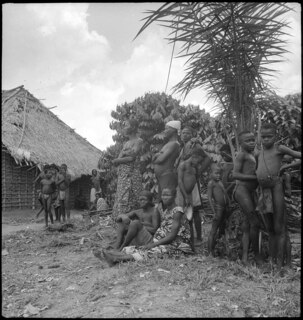 Belgisch-Kongo, Molanda (Malanda): Menschen; Mehrere Personen stehen und sitzen neben Pflanzen und einer Strohhütte
