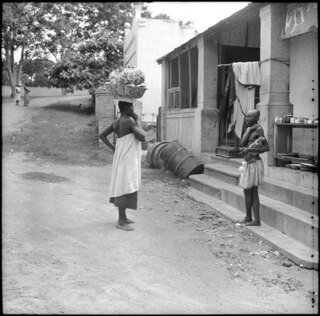 Belgisch-Kongo, Lisala: Dorfladen; Barfüssige Frau mit Korb auf dem Kopf und barfüssiger Mann stehen vor einem Dorfladen