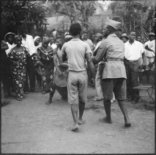 Belgisch-Kongo, Leopoldville (Kinshasa): Strassenszene; Gruppe von Personen auf einer Strasse bildet einen Kreis und tanzt zu Trommelklängen des Tam-Tam, darunter ein uniformierter Polizist
