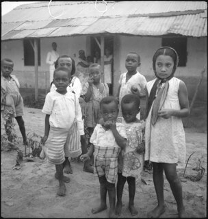 Belgisch-Kongo, Thysville (Mbanza Ngungu): Kinder; Mehrere frabige Kinder stehen barfuss aber bekleidet vor einer Hütte mit Wellblechdach im Stadtteil "Belge"