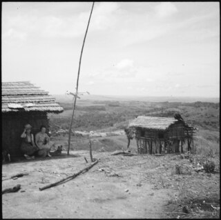 Belgisch-Kongo, Thysville (Mbanza Ngungu): Hütte; Kleine Strohhütte auf Pfählen (für Vorräte?) auf einer Anhöhe mit Blick auf eine Ebene daneben eine weitere Hütte mit zwei Personen im Schatten davor kauernd