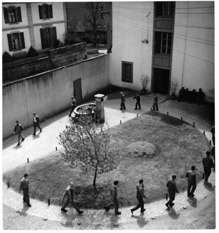 Kolonne von Gefangenen beim Spazieren im Innenhof mit Grünanlage und Brunnen, rechts vier Männer auf einer Bank sitzend