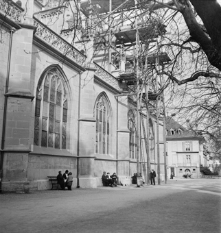 Südfassade des Münster-Kirchenschiffes, davor Sitzbänke mit Menschen, zwei Kinderwagen, Baugerüst aus Holz. Im Hintergrund zwei Kinder beim Löschbrunnen