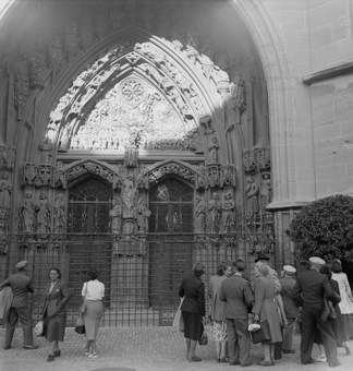 Hauptportal Münster mit Lichteinfall (Abendsonne) auf das Jüngste Gericht. Davor eine Besuchergruppe: Damen in Kleidern und mit Handtaschen, die Herren in Anzügen und Mützen oder Hüten