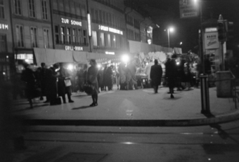 Bärenplatz am Abend, Restaurants und ihre Leuchtschriften: Zur Sonne, Gfeller-Rindlisbacher,  Marktstände,Verkehrsampel, Bus-Haltestelle (Bus Nr. 6, Tram Nr. 4), Telefonkabine mit Leuchtschrift und Reklameplakat («Radio Steiner»). Zeitungsverkäufer, Marktbesucher und Passanten