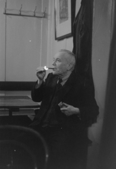 Älterer Mann mit Zigarre und Zündholzschachtel auf Eckbank in Restaurant. An der Wand Garderobe mit 4 Haken, Bild und Mantel