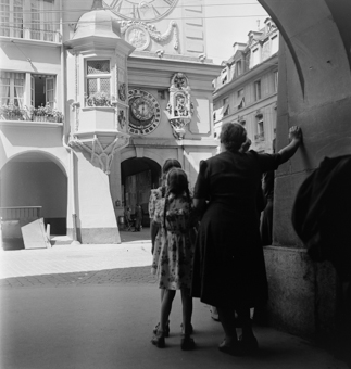 Zeitglockenturm mit Glockenspiel und astronomischer Uhr, Erker. Frau und zwei Mädchen mit Zöpfen und Sommerkleidern schauen zum Glockenspiel. Im Turmdurchgang Plakatwand, davor Mann auf Klappstuhl und zwei Kinder