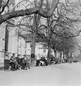 Münsterplattform mit Menschen auf Sitzbänken an der Frühlingssonne. Kinder in Schürzen auf Bank beim Sandkasten, Kind auf Dreiradvelo. Einige Frauen am Stricken