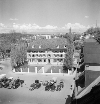Polizeigebäude, Sitz von Kantonspolizei und Stadtpolizei (Schild «Stadtpolizei»). Fahrräder vor dem Gebäude. Zahlreiche parkierte Autos und Lastwagen auf dem Waisenhausplatz wegen eines Anlasses. Zwei Polizisten mit Fahrrädern. Aufnahme aus dem Dachfenster eines Hauses am Waisenhausplatz (Seite Holländerturm)