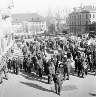 Waisenhausplatz mit Kleintier-Markt, Kisten und Käfigen (Hühner, Kaninchen) und Holz-Markt (Stühle, Bottiche etc). Marktbesucher und Händler, Automobile und Lastwagen. Im Hintergrund links das Polizeigebäude im ehemaligen Waisenhaus, rechts das Gebäude der Neuen Mädchenschule (Gründungsjahr 1851)