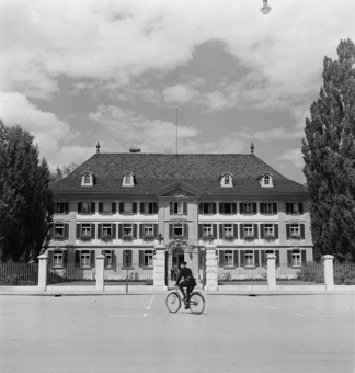 Polizist auf Fahrrad vor dem Polizeigebäude (ehemaliges Waisenhaus). Schild «Stadtpolizei», Fahrräder vor dem Gebäude, drei Männer am Verlassen des Gebäudes