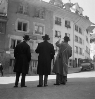 Haus Nydeggstalden/Läuferplatz mit Einschussloch vom «Stäcklichrieg» 1802. Davor vier Männer mit Mänteln und Hüten beim Betrachten des Hauses. Rechts Nydeggstalden mit parkiertem Auto