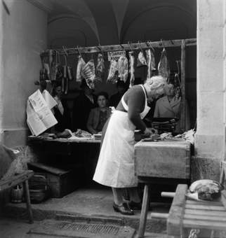 Stand von Fr. Gäumann Wichtrach an der Kesslergasse (1967 umbenennt in Münstergasse). Fleischverkäuferin in weisser Schürze am Schneiden auf dem Metzger-Block. Fleischstücke und Würste an Haken, auf der Theke ein Schinken. Waage, Zeitungspapier zum Einpacken der Ware. Einige Kundinnen am Warten