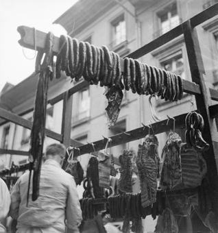 Fleischstand mit Fleischhändler. Würste und Fleischstücke an Haken. Im Hintergrund Häuser der Kesslergasse (heute Münstergasse).