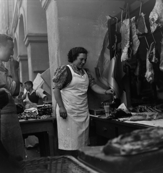 Stand mit Fleischhändlern an der Kesslergasse (1967 umbenennt in Münstergasse), Metzgerschürze, Verkäuferinnenschürze. Fleischstücke an Haken und auf der Theke. Waage mit Wägesteinen. 2 Gläser mit Tee oder Kaffee