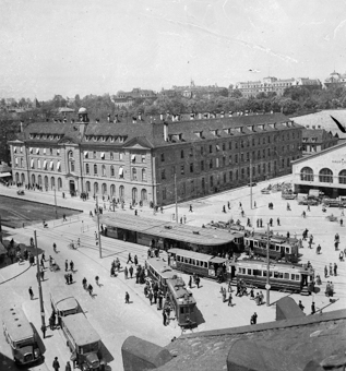 Bahnhofplatz mit Tramhaltestelle, mehrere Trams, Saurer-Busse von 1929 und 1910 mit Anhänger, Fussgänger. In der Mitte der Burgerspittel, rechts das Bahnhofsgebäude, davor Autos und Lieferwagen. Im Hintergrund Obergericht, Häuser Länggassquartier und Universität. Bild aufgenommen aus Dachfenster des Warenhauses Loeb