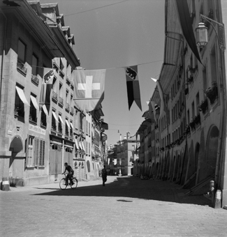 Haus Junkerngasse 58 mit Schild «Lager in fertigen Betten» (heute Utiger Bettenhaus). Junger Mann mit Fahrrad, Mann mit Rucksack. Im Hintergrund derJunkerngassbrunnen und das Haus Ecke Junkerngasse/Bubenbergrain, diverse Flaggen hangen quer über der Gasse