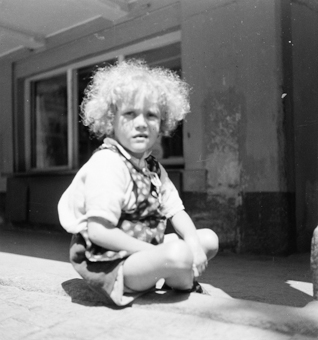 Sitzendes Kind mit blonden Locken und Sommerkleidung, im Hintergrund Laube mit Schaufenster