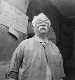 Mann in Arbeitsschürze, Laubenbogen mit Schild «Ankauf Lumpen & Zeitungen, Bücher & Metallabfälle, Räumen von Estrichen»