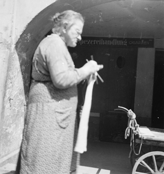 Frau mit Strickarbeit  im Stehen, Laubenbogen mit Holzleiterwagen. In der Laube Schild «Spezereihandlung, Cigarren»