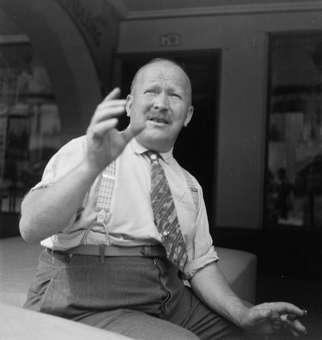 Mann mit Krawatte und Hosenträgern, hinten Laube mit Eingangstüre Nr 70 und Schaufenster